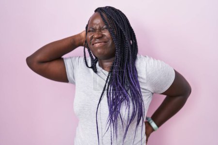 Foto de Mujer africana joven de pie sobre fondo rosa sufriendo de dolor de cuello lesión, tocando el cuello con la mano, dolor muscular - Imagen libre de derechos