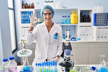 Photo for Brunette woman working at scientist laboratory smiling positive doing ok sign with hand and fingers. successful expression. - Royalty Free Image