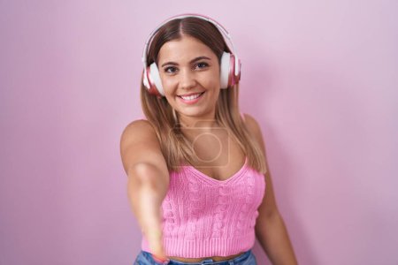 Foto de Mujer rubia joven escuchando música usando auriculares que sonríen amigables ofreciendo un apretón de manos como saludo y bienvenida. negocios exitosos. - Imagen libre de derechos