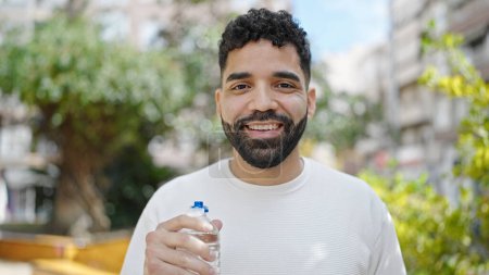 Téléchargez les photos : Jeune homme hispanique souriant confiant tenant bouteille d'eau au parc - en image libre de droit