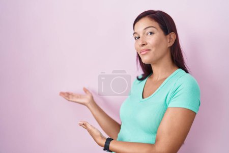 Foto de Mujer hispana joven de pie sobre fondo rosa invitando a entrar sonriendo natural con la mano abierta - Imagen libre de derechos