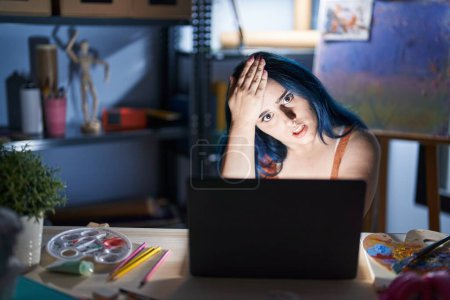 Jeune fille moderne avec les cheveux bleus assis au studio d'art avec ordinateur portable la nuit surpris avec la main sur la tête pour erreur, rappelez-vous erreur. oublié, mauvais concept de mémoire. 