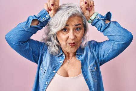 Téléchargez les photos : Femme d'âge moyen avec les cheveux gris debout sur fond rose faisant geste drôle avec doigt sur la tête comme cornes de taureau - en image libre de droit