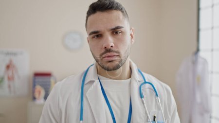 Téléchargez les photos : Jeune homme hispanique médecin debout avec une expression sérieuse à la clinique - en image libre de droit