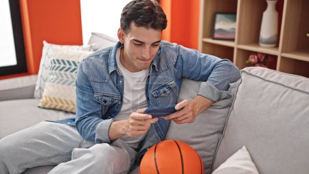 Téléchargez les photos : Jeune homme hispanique regardant le jeu de basket sur smartphone à la maison - en image libre de droit