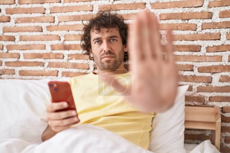 Photo for Hispanic young man using smartphone on the bed with open hand doing stop sign with serious and confident expression, defense gesture - Royalty Free Image