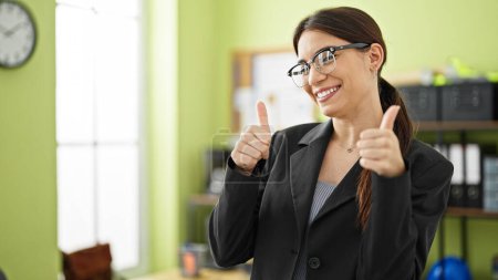 Photo for Young beautiful hispanic woman business worker doing thumbs up gesture smiling at office - Royalty Free Image