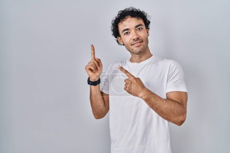Foto de Hombre hispano parado sobre un fondo aislado sonriendo y mirando a la cámara apuntando con dos manos y dedos hacia un lado. - Imagen libre de derechos