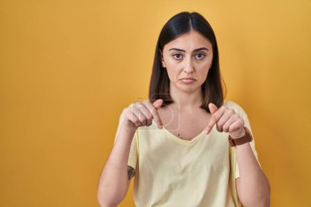 Foto de Chica hispana con camiseta casual sobre fondo amarillo apuntando hacia abajo mirando triste y molesto, indicando la dirección con los dedos, infeliz y deprimido. - Imagen libre de derechos