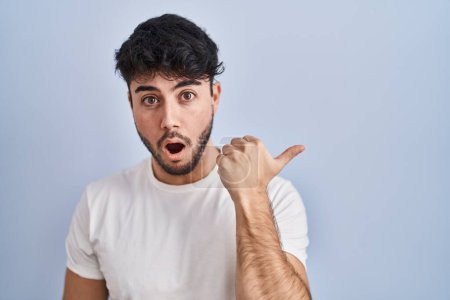 Foto de Hispanic man with beard standing over white background surprised pointing with hand finger to the side, open mouth amazed expression. - Imagen libre de derechos