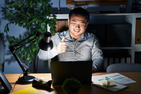 Foto de Joven hombre chino que trabaja usando computadora portátil en la noche haciendo un gesto feliz pulgares hacia arriba con la mano. aprobando la expresión mirando a la cámara mostrando éxito. - Imagen libre de derechos