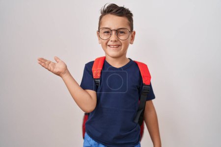 Foto de Pequeño niño hispano con gafas y mochila de estudiante celebrando el logro con sonrisa feliz y expresión ganadora con la mano levantada - Imagen libre de derechos