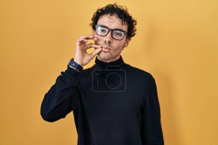 Foto de Hombre hispano de pie sobre fondo amarillo boca y labios cerrados como cremallera con los dedos. secreto y silencioso, tabú hablando - Imagen libre de derechos