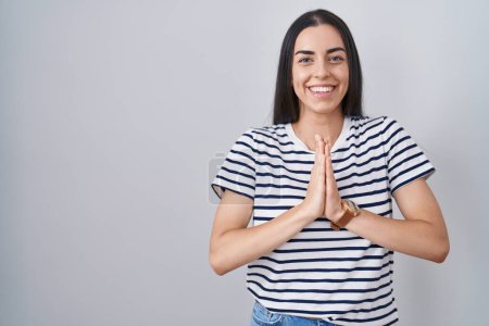 Foto de Joven morena vistiendo camiseta a rayas rezando con las manos juntas pidiendo perdón sonriendo confiada. - Imagen libre de derechos