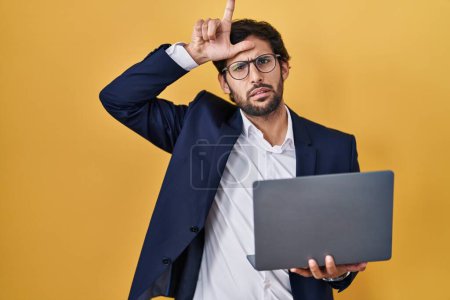 Téléchargez les photos : Beau latin homme travaillant à l'aide d'un ordinateur portable se moquant des gens avec les doigts sur le front faisant geste perdant moquerie et insultant. - en image libre de droit