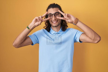 Photo for Young hispanic man standing over yellow background doing peace symbol with fingers over face, smiling cheerful showing victory - Royalty Free Image
