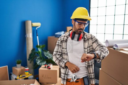 Photo for Young hispanic man with beard working at home renovation checking the time on wrist watch, relaxed and confident - Royalty Free Image