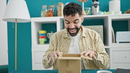 Foto de Joven hombre hispano desempacando caja de cartón con expresión sorpresa en el comedor - Imagen libre de derechos