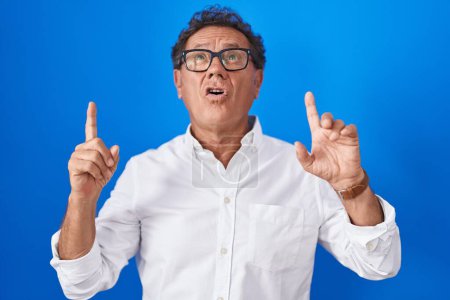 Foto de Hombre hispano de mediana edad de pie sobre fondo azul asombrado y sorprendido mirando hacia arriba y señalando con los dedos y los brazos levantados. - Imagen libre de derechos