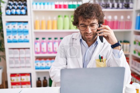 Téléchargez les photos : Young hispanic man pharmacist talking on smartphone using laptop at pharmacy - en image libre de droit