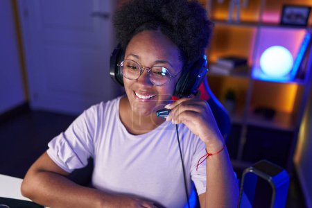 Foto de Mujer afroamericana streamer sonriendo confiado sentado en la mesa en sala de juegos - Imagen libre de derechos