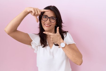 Téléchargez les photos : Femme hispanique du Moyen Âge portant T-shirt blanc décontracté et lunettes souriantes faisant cadre avec les mains et les doigts avec le visage heureux. concept de créativité et photographie. - en image libre de droit