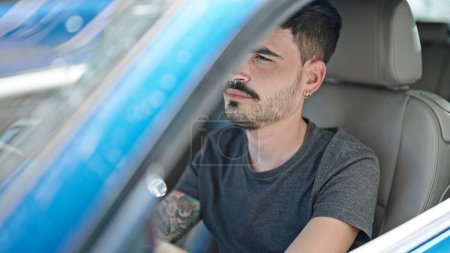 Foto de Joven hombre hispano conduciendo coche con expresión relajada en la calle - Imagen libre de derechos