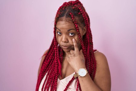 Foto de Mujer afroamericana con cabello trenzado de pie sobre fondo rosa apuntando al ojo observándote gesto, expresión sospechosa - Imagen libre de derechos