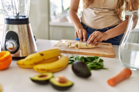 Foto de Joven hermosa mujer hispana preparando batido de verduras con licuadora cortando plátano en la cocina - Imagen libre de derechos