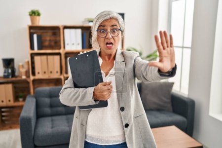Téléchargez les photos : Femme d'âge moyen avec les cheveux gris au bureau de consultation faire arrêter geste avec les paumes des mains, expression de colère et de frustration - en image libre de droit