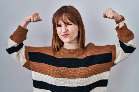 Foto de Mujer hermosa joven con suéter a rayas sobre un fondo aislado que muestra los músculos de los brazos sonriendo orgulloso. concepto de fitness. - Imagen libre de derechos