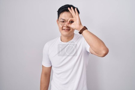 Téléchargez les photos : Jeune homme asiatique debout sur fond blanc faisant un geste correct avec la main souriante, les yeux regardant à travers les doigts avec un visage heureux. - en image libre de droit