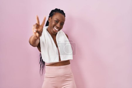 Téléchargez les photos : Belle femme noire portant des vêtements de sport et une serviette sur fond rose souriant en regardant la caméra montrant des doigts faisant signe de victoire. numéro deux. - en image libre de droit