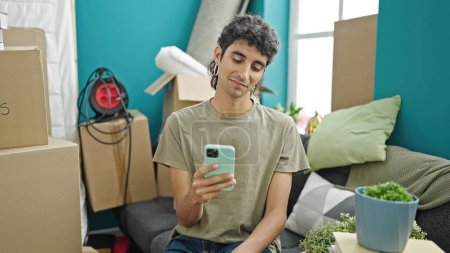 Foto de Hombre hispano joven usando teléfono inteligente sentado en un sofá en un nuevo hogar - Imagen libre de derechos