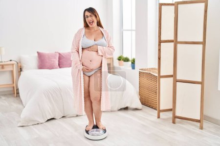 Téléchargez les photos : Femme enceinte sur la machine de poids clin d'oeil en regardant la caméra avec une expression sexy, gai et heureux visage. - en image libre de droit