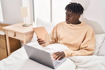 Photo for African american man reading book using laptop at bedroom - Royalty Free Image