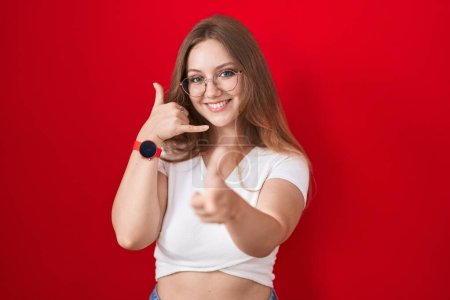 Téléchargez les photos : Jeune femme caucasienne debout sur fond rouge souriant faire parler au téléphone geste et vous pointant du doigt. Appelle-moi. - en image libre de droit