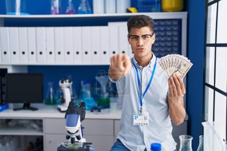 Foto de Joven hispano trabajando en laboratorio científico sosteniendo dólares señalando con el dedo a la cámara y a ti, gesto confiado que parece serio - Imagen libre de derechos