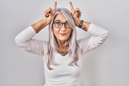 Téléchargez les photos : Femme d'âge moyen avec les cheveux gris debout sur fond blanc faisant un geste drôle avec le doigt sur la tête comme cornes de taureau - en image libre de droit