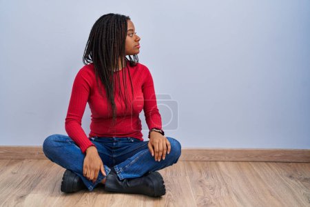 Téléchargez les photos : Jeune afro-américain avec des tresses assises sur le sol à la maison regardant vers le côté, pose de profil relax avec le visage naturel et sourire confiant. - en image libre de droit