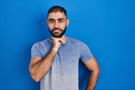 Photo for Middle east man with beard standing over blue background with hand on chin thinking about question, pensive expression. smiling with thoughtful face. doubt concept. - Royalty Free Image