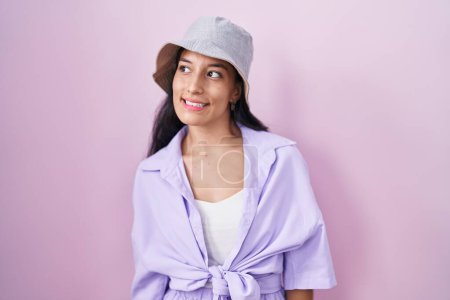 Téléchargez les photos : Jeune femme hispanique debout sur fond rose portant chapeau regardant de côté avec le sourire sur le visage, expression naturelle. riant confiant. - en image libre de droit
