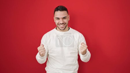 Téléchargez les photos : Jeune homme hispanique souriant confiant debout avec geste gagnant sur fond rouge isolé - en image libre de droit