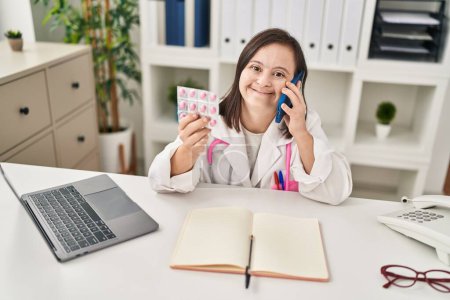 Téléchargez les photos : Femme trisomique portant un uniforme de médecin parlant sur le smartphone tenant des pilules contraceptives à la clinique - en image libre de droit