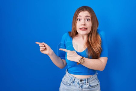 Photo for Redhead woman standing over blue background pointing aside worried and nervous with both hands, concerned and surprised expression - Royalty Free Image