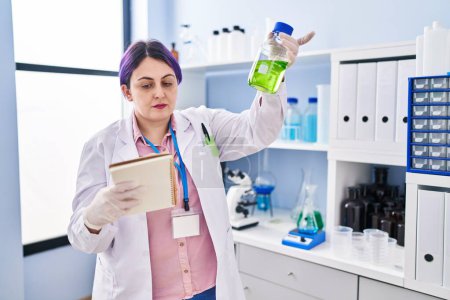 Photo for Young beautiful plus size woman scientist reading notebook holding bottle with liquid at laboratory - Royalty Free Image