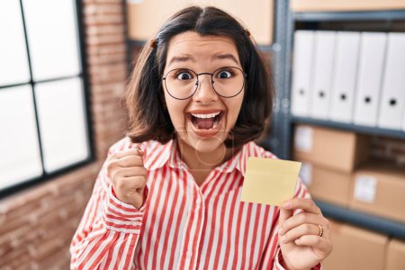 Photo for Young hispanic woman holding paper reminder at the office screaming proud, celebrating victory and success very excited with raised arms - Royalty Free Image
