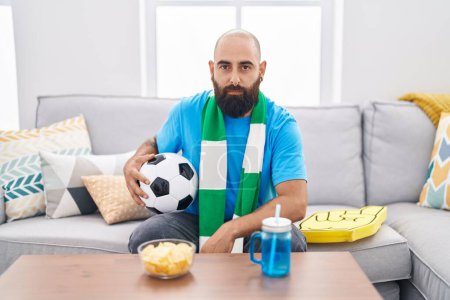 Photo for Young hispanic man with beard and tattoos football hooligan holding ball supporting team thinking attitude and sober expression looking self confident - Royalty Free Image