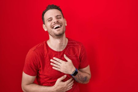 Téléchargez les photos : Jeune homme hispanique debout sur fond rouge souriant et riant fort à haute voix parce que drôle blague folle avec les mains sur le corps. - en image libre de droit