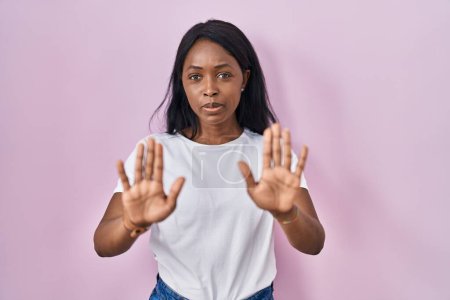 Foto de Mujer joven africana con camiseta blanca casual alejando las palmas de las manos mostrando rechazo y negación con miedo y expresión repugnante. parada y prohibido. - Imagen libre de derechos